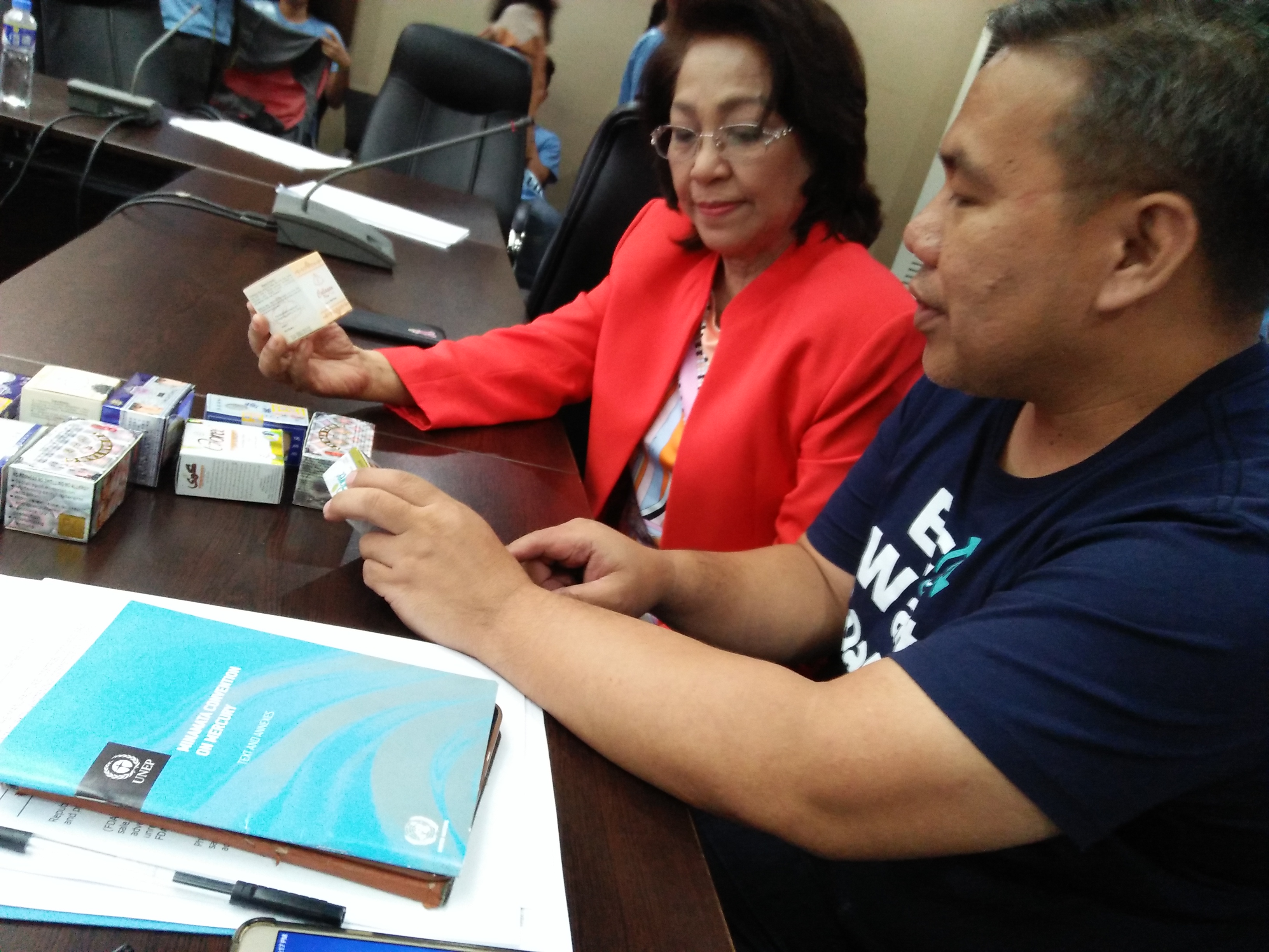 EcoWaste Coalition's Chemical Safety Campaigner Thony Dizon show Councilor Elizabeth Delarmente samples of dangerous skin lightening products containing mercury, a toxic chemical that is not allowed in cosmetic product formulations under the ASEAN Cosmetic Directive.  The Minamata Convention on Mercury (the blue book at the foreground) has set a phase-out by 2020 of skin lightening creams and soaps with mercury above 1 ppm. 