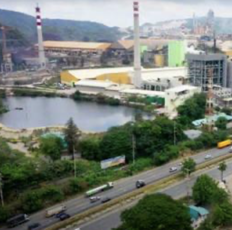 ऊँची चिमनियों और बड़ी इमारतों वाला एक औद्योगिक परिसर पानी के एक बड़े भंडार के बगल में स्थित है, जिसमें आगे एक राजमार्ग और कई वाहन हैं, और पृष्ठभूमि में पहाड़ हैं।.