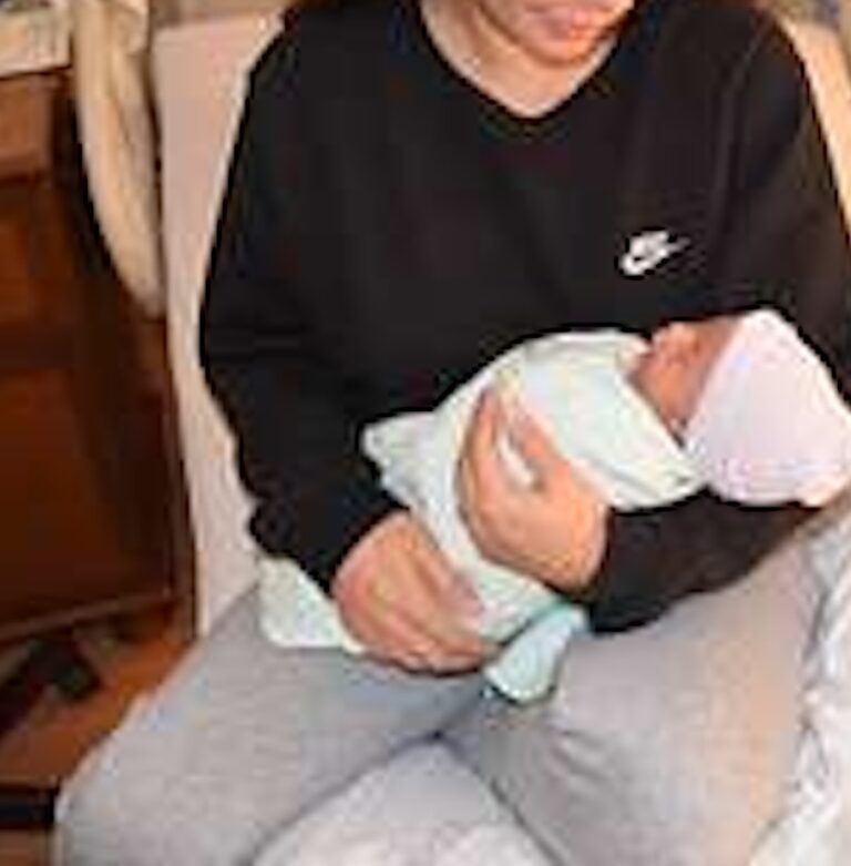 A person wearing a black Nike sweatshirt holds a baby wrapped in a white blanket and hat while sitting in a chair indoors.