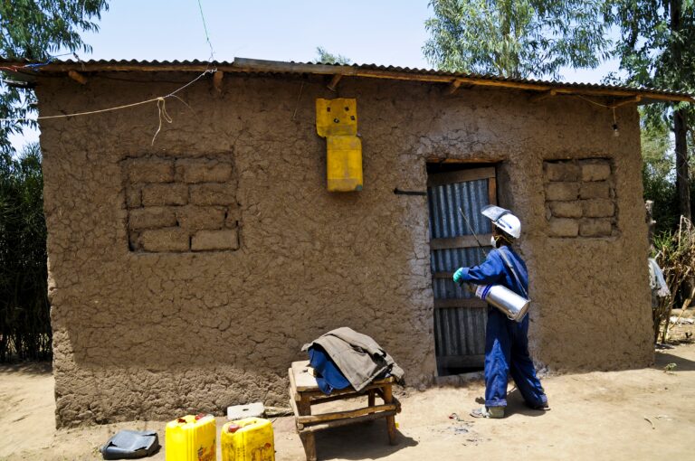 A person in protective clothing sprays disinfectant on the door of a small, mud-brick house with a tin roof. Several yellow containers and a wooden bench are positioned in front of the building.