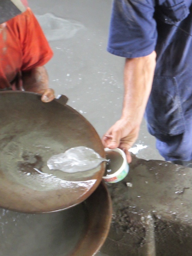 Two people use a large, shallow pan to separate material in water; one person pours liquid from the pan into a small cup held by the other person. Their faces are not visible.