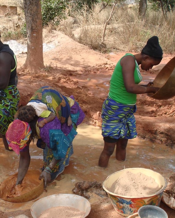 Two women stand in muddy water outdoors, wearing colorful skirts. One bends over, washing something, while the other uses a large shallow pan to sift material. Bowls and basins are on the ground around them.