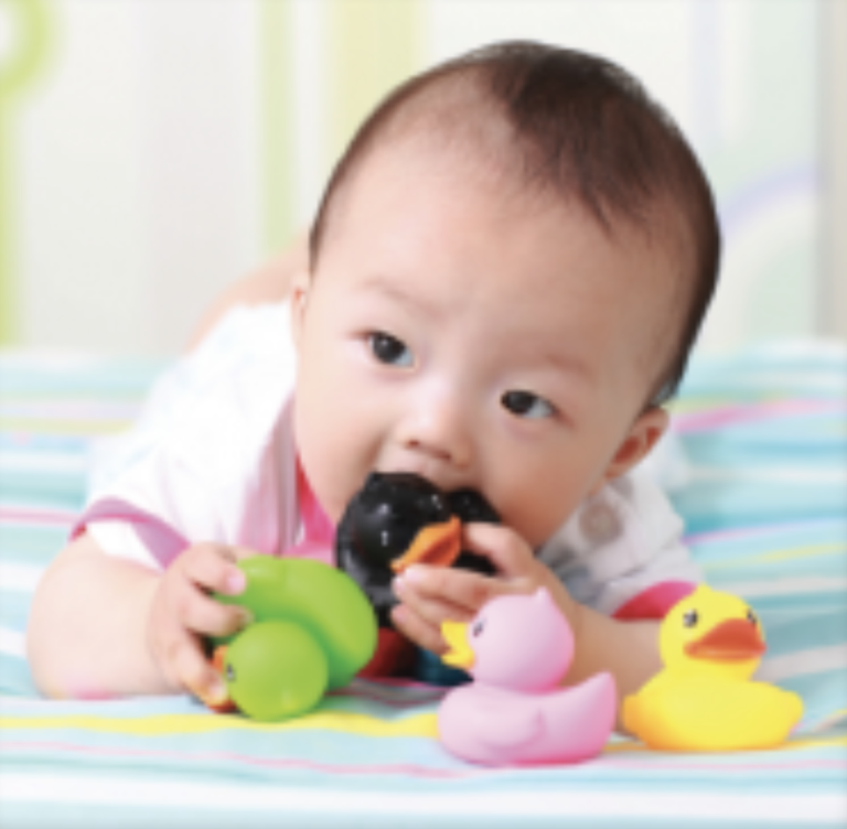 A baby lies on a striped blanket, holding green and pink rubber duck toys, and chewing on a black rubber duck. A yellow rubber duck is also in front of the baby.