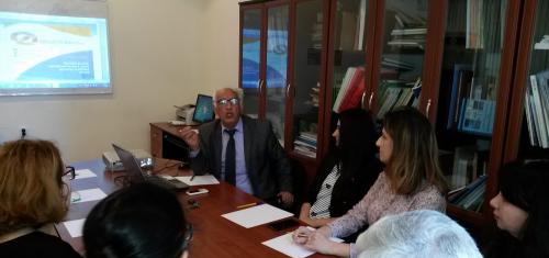 A man in a suit speaks to a group of people seated around a table in an office setting. There are papers on the table, a laptop, and a presentation projected on the wall. Bookshelves with files are in the background.