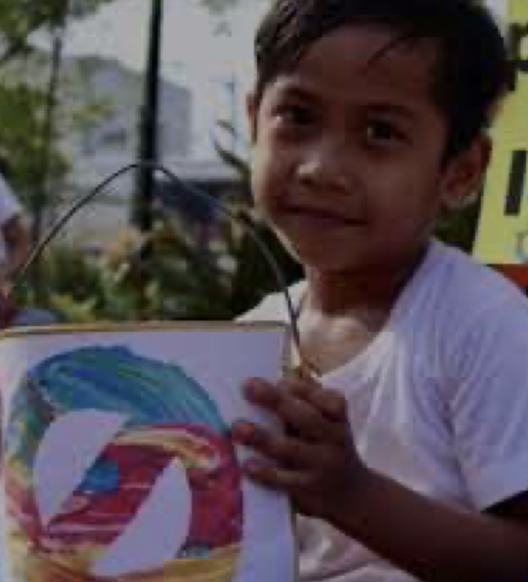 A young boy wearing a white shirt holds up a drawing colored with red, yellow, blue, and green. The drawing is attached to a small handle, and the boy is outdoors with blurred greenery and buildings in the background.