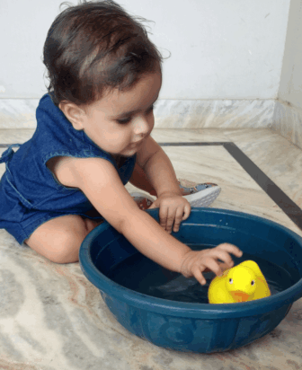 A toddler in a blue outfit sits on a marble floor, reaching into a blue plastic basin filled with water and playing with a yellow rubber duck.