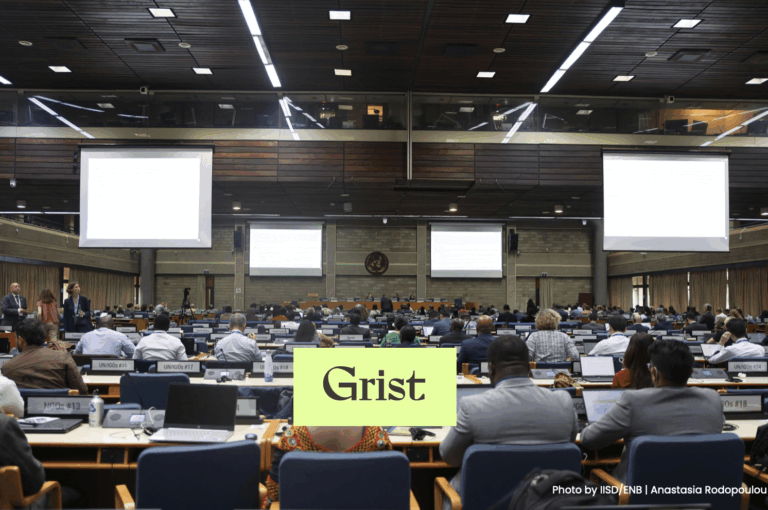 A large conference room filled with people seated at rows of desks, facing the front where projection screens are displayed. The word Grist is overlaid in a green box near the center of the image.