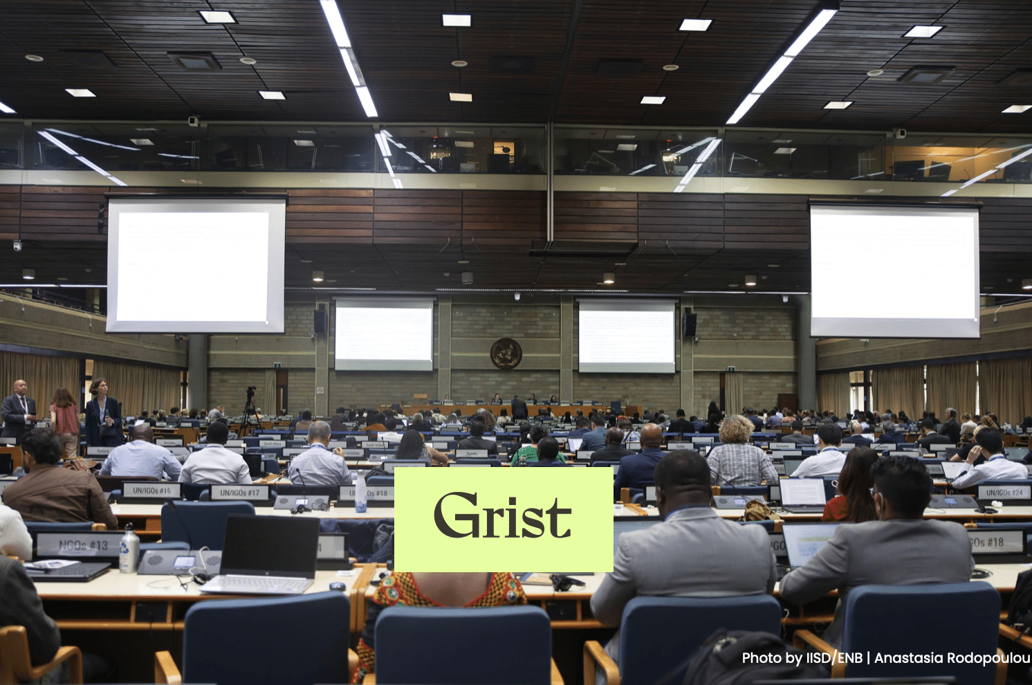 A large conference room filled with people seated at rows of desks, facing the front where projection screens are displayed. The word Grist is overlaid in a green box near the center of the image.