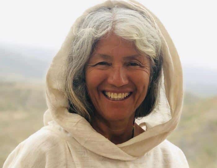 A woman with gray and black hair, wearing a light-colored hooded garment, smiles at the camera with a natural outdoor background behind her.