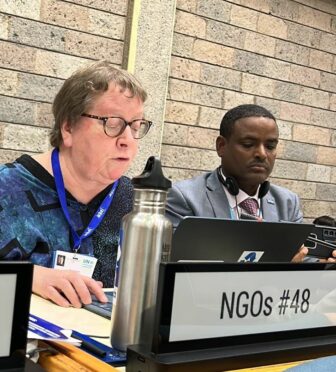 Two people sit at a conference table labeled NGOs #48. The woman on the left is speaking, and the man on the right is looking at a tablet. Both wear badges and are in front of a brick wall. A metal water bottle is visible.