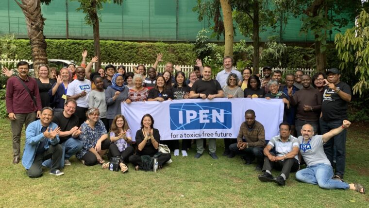 A large group of people pose outdoors on grass, some seated and some standing, behind a banner that reads “IPEN for a toxics-free future.” There are trees and a fence in the background.