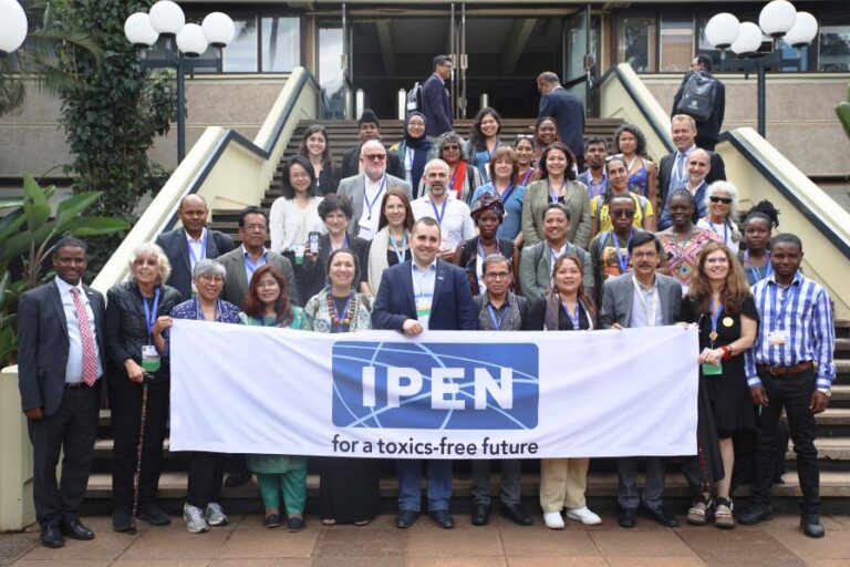 A large group of people pose for a photo on steps outside a building, holding a banner that reads IPEN for a toxics-free future. The group appears diverse in age, gender, and ethnicity.