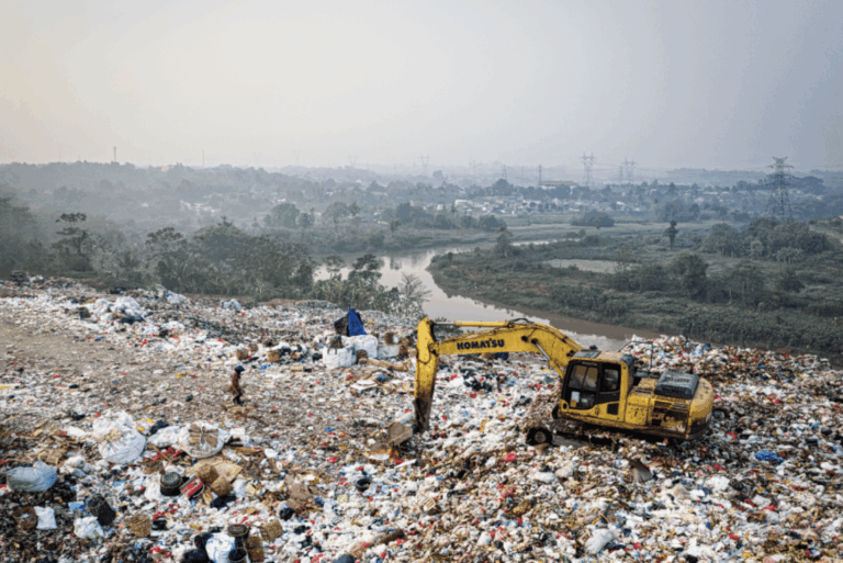 A yellow excavator sits atop a large landfill covered with assorted waste. A person walks nearby. In the background, greenery, a river, and distant buildings are visible under a hazy sky.