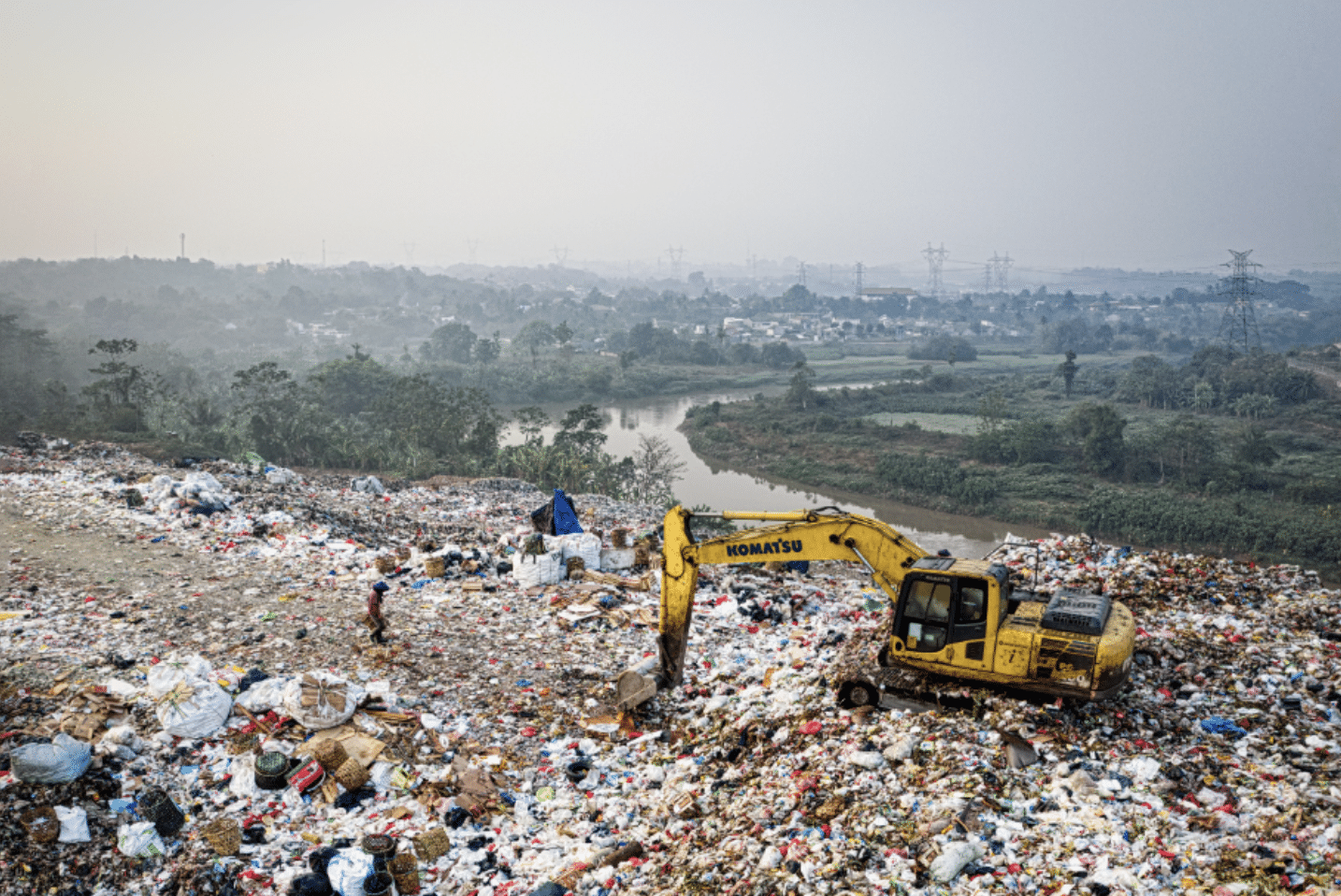 A yellow excavator sits atop a large landfill covered with assorted waste. A person walks nearby. In the background, greenery, a river, and distant buildings are visible under a hazy sky.