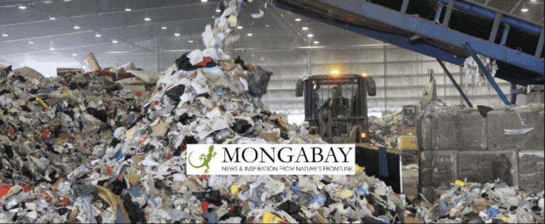 A large pile of mixed waste is being dumped in a recycling facility. A bulldozer is visible behind the pile, and an industrial conveyor belt is seen on the right. The Mongabay logo is overlaid on the image.