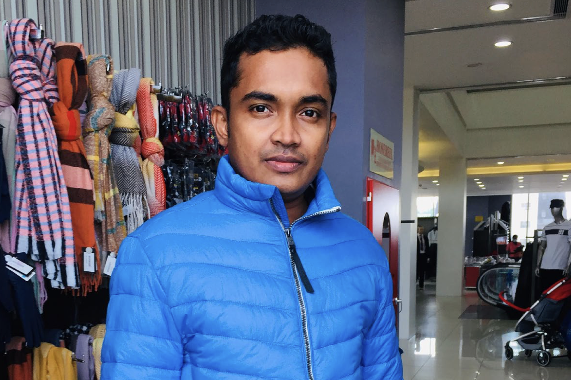 A man wearing a bright blue puffer jacket stands indoors near a display of scarves and clothing accessories. The background shows a store interior with mannequins, shelves, and escalators.