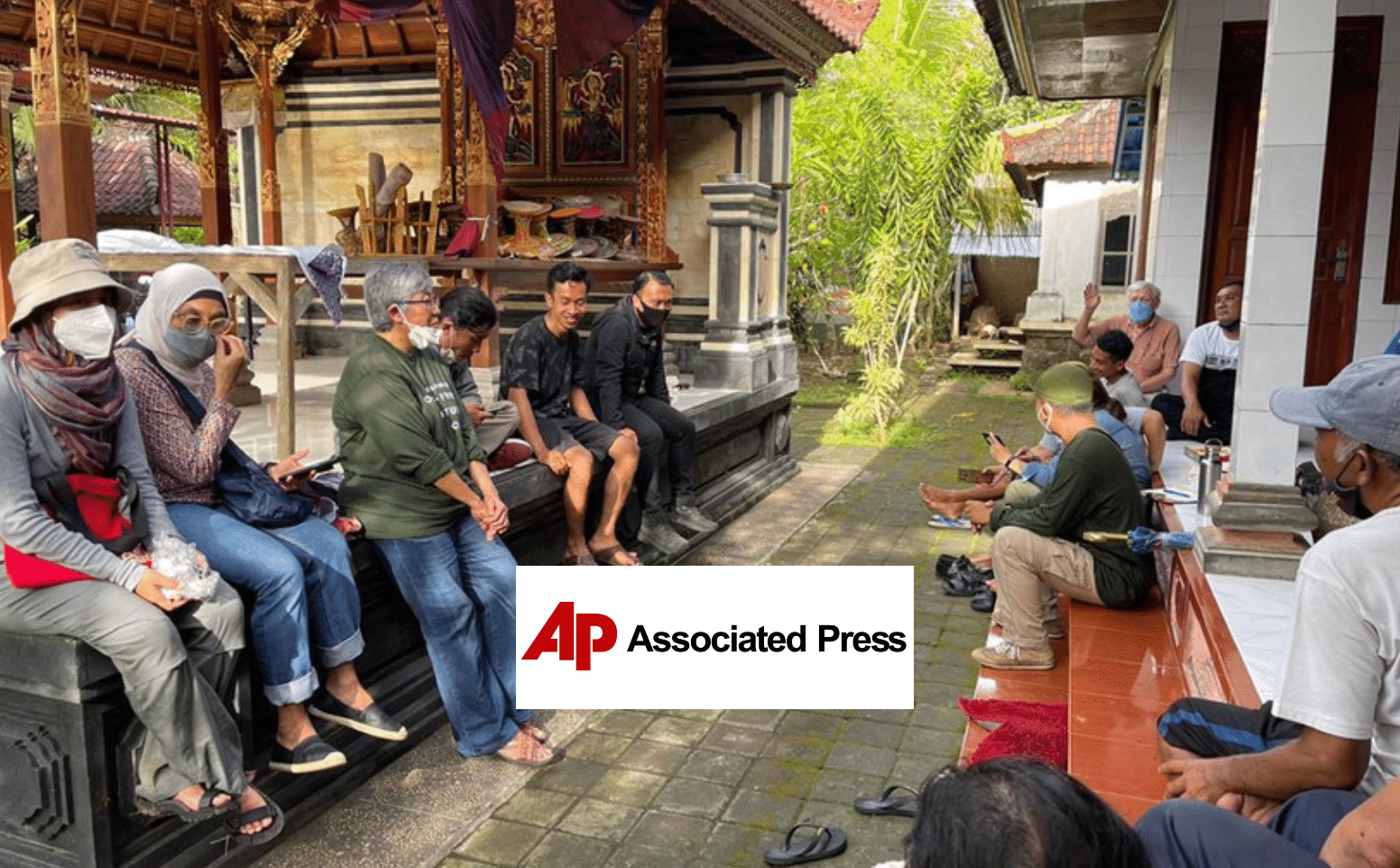 A group of people sit together outdoors in a courtyard area, some wearing masks and headscarves. They appear to be in conversation or using phones. The AP Associated Press logo is overlaid at the bottom center.