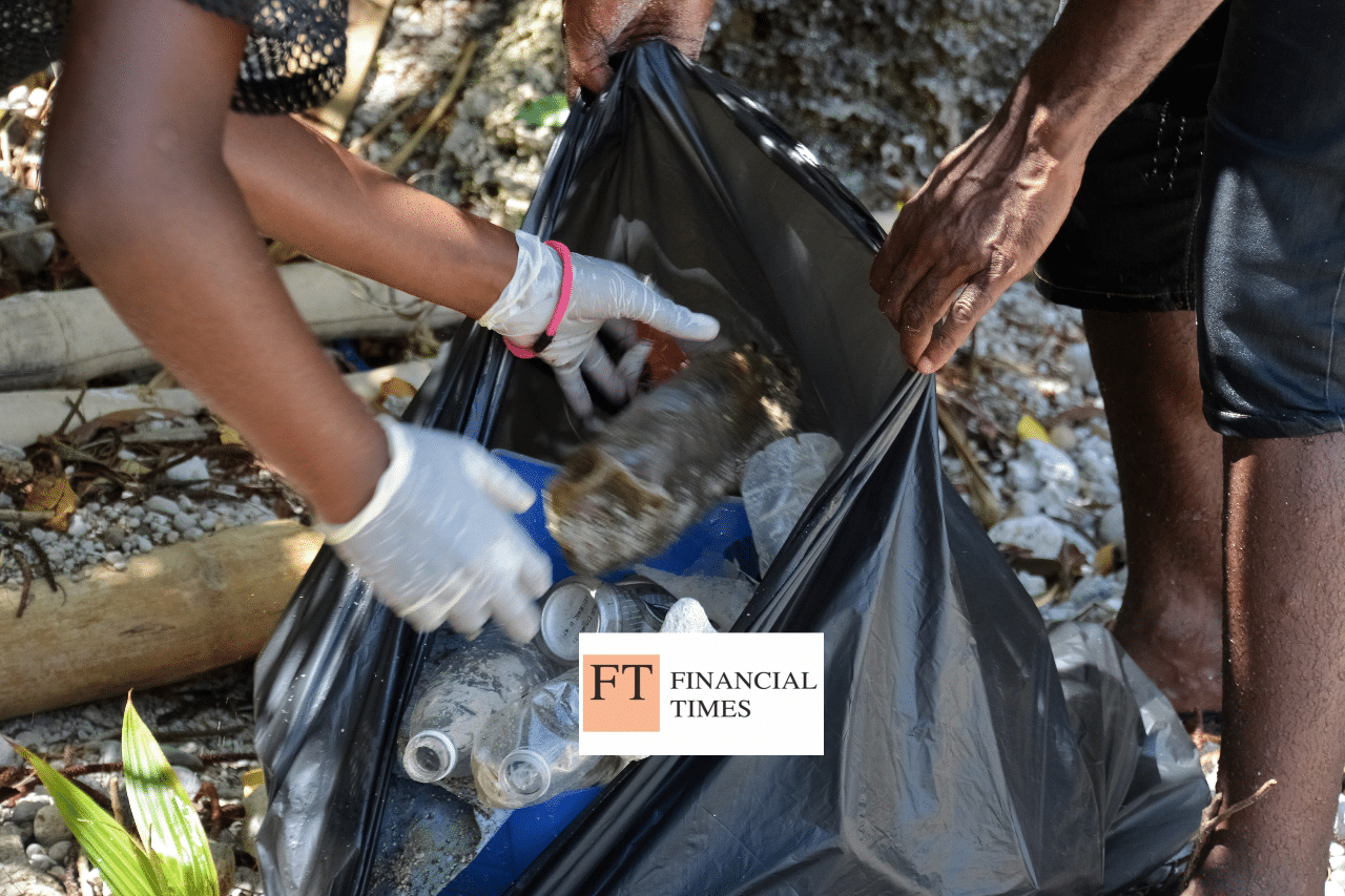 Two people wearing gloves place plastic bottles and other trash into a large black garbage bag during a cleanup; the Financial Times logo is overlaid on the image.
