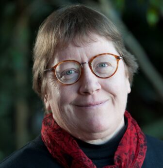 A person with short light brown hair, wearing round glasses, a black top, and a red patterned scarf, is smiling gently at the camera. The background is softly blurred greenery.