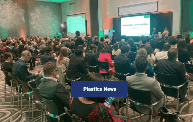 A large group of people sit in rows facing a stage with two screens displaying presentation slides in a conference room. The overlay text reads “Plastics News.”.