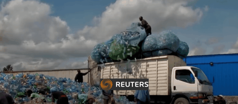 Several people load large bags filled with plastic bottles onto a white truck surrounded by piles of plastic waste, under a partly cloudy sky. The Reuters logo is displayed at the bottom center of the image.