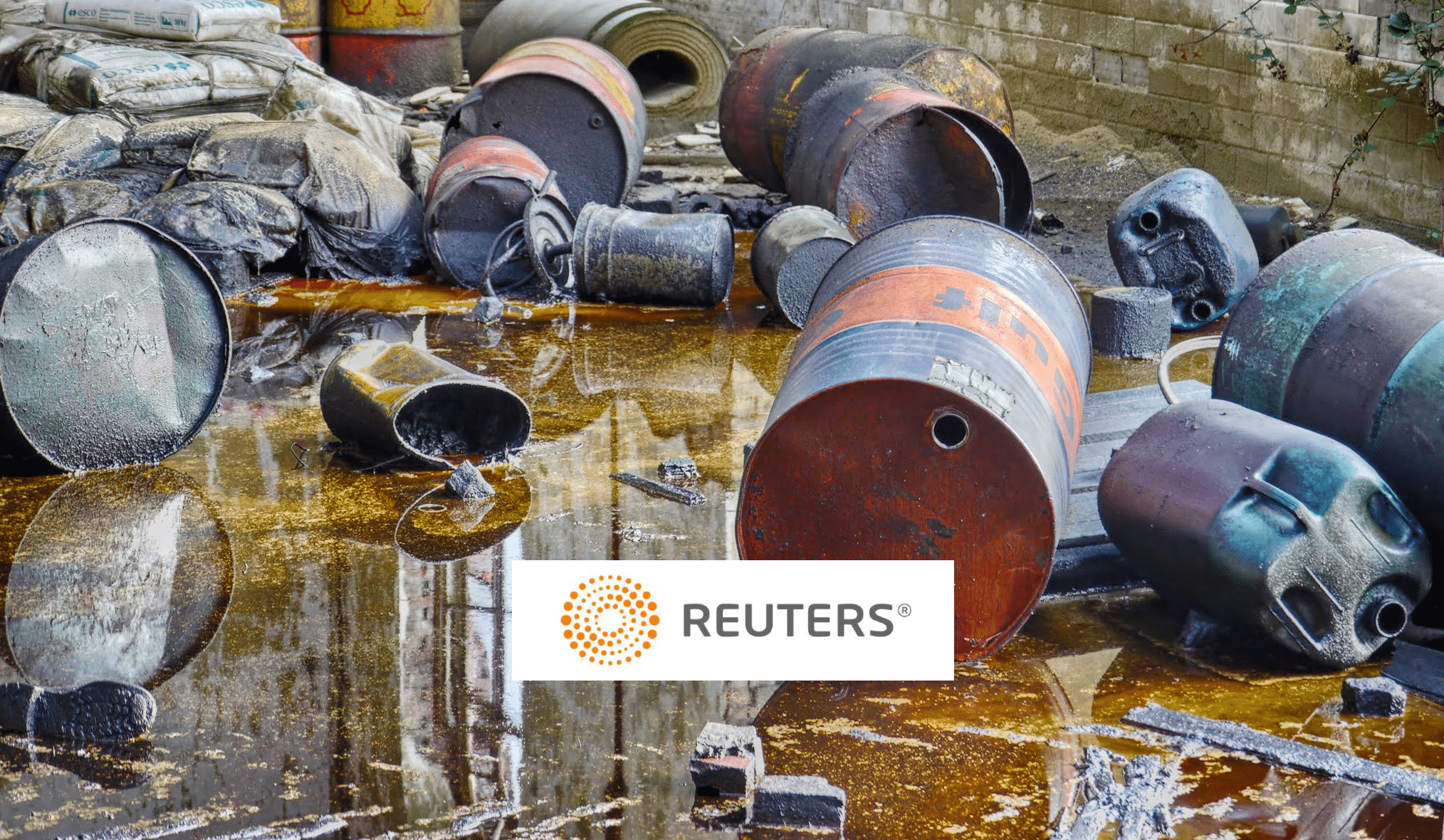 Rusty barrels and containers are scattered on the ground, leaking oily liquid. Worn sacks and industrial debris are visible in the background. A Reuters logo is overlaid on the image.