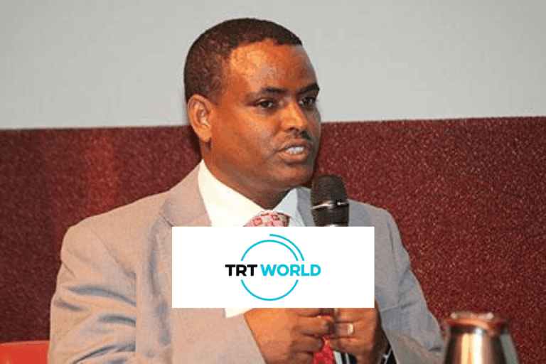 A man in a light gray suit and red tie speaks into a microphone while seated. The background is plain, and a TRT World logo is overlaid on the image, partially covering his hands.
