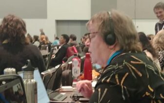 A person wearing a headset speaks into a microphone at a conference, sitting among other attendees, with water bottles and laptops on the table.