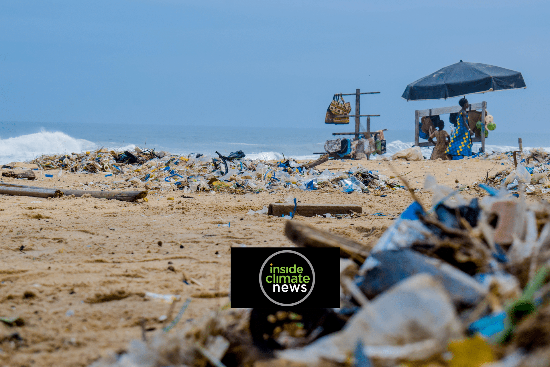 Plastic and other debris litter a sandy beach, with a few people and a black umbrella in the background near the ocean. The sky is overcast. The Inside Climate News logo appears at the bottom center.