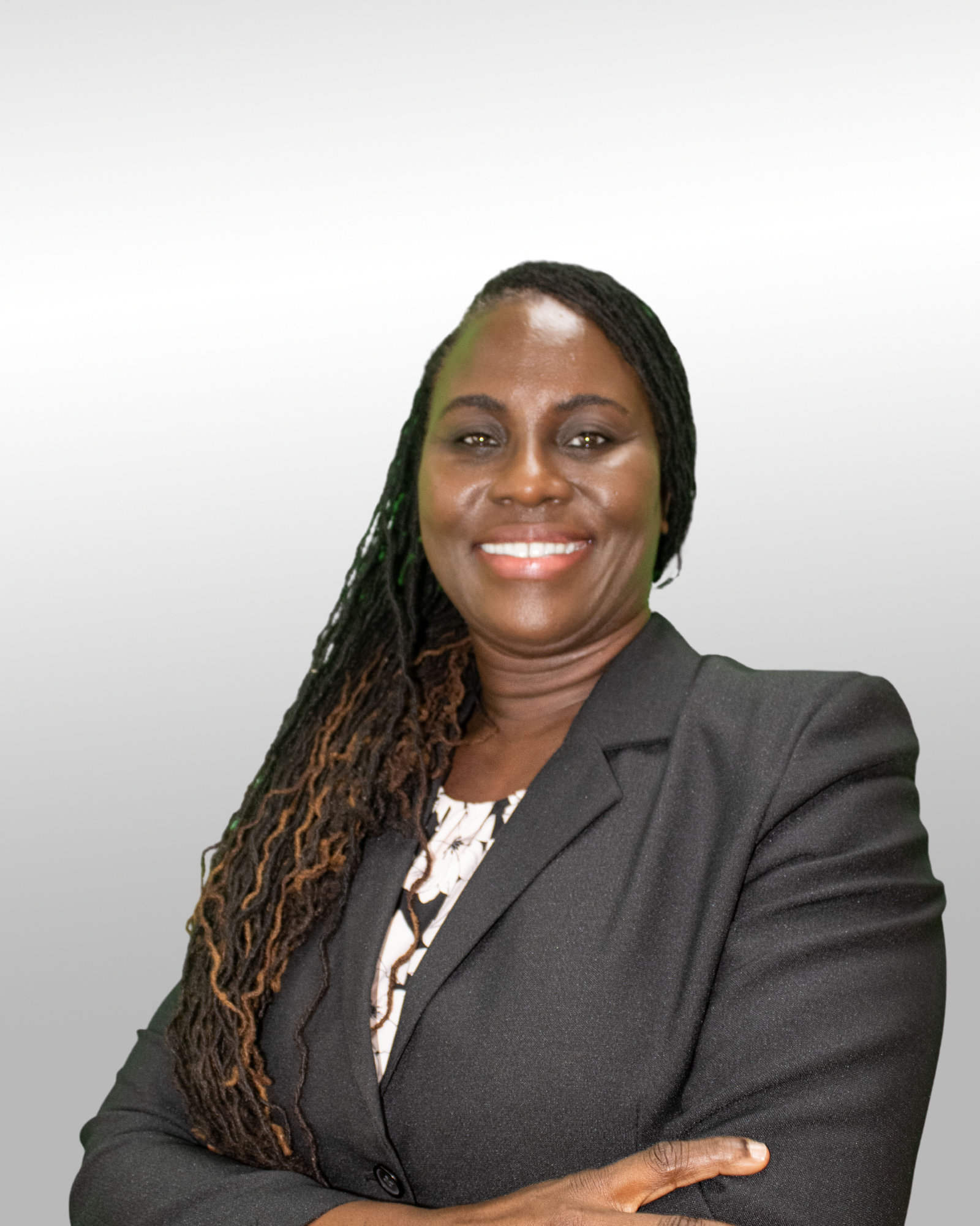 A woman with long, braided hair wearing a black blazer and patterned top stands against a plain light gray background, smiling with her arms crossed.