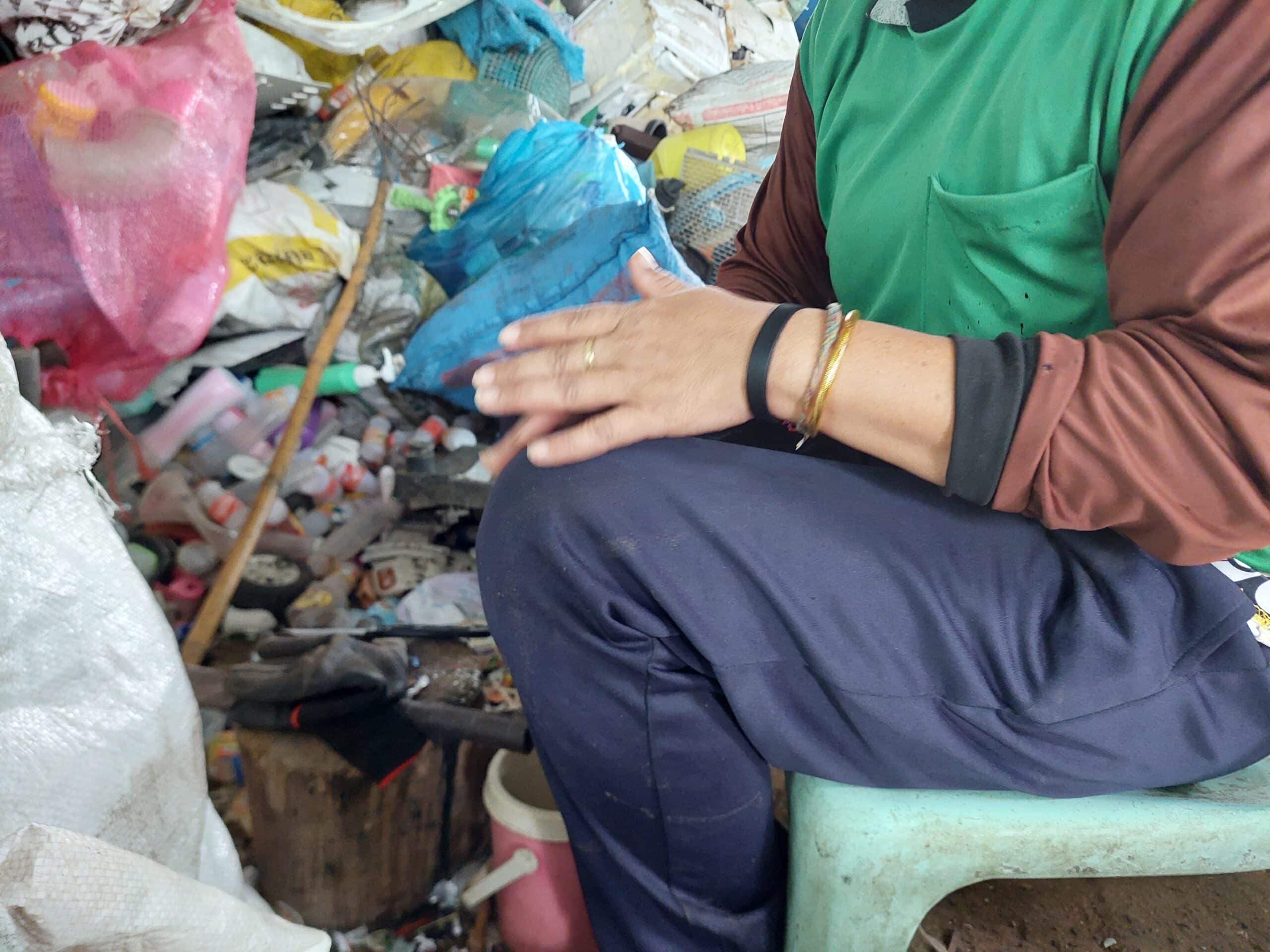 A person sits on a small stool in a cluttered area filled with bags, plastic bottles, and various recyclables, with hands resting on their knees and only their torso and hands visible.