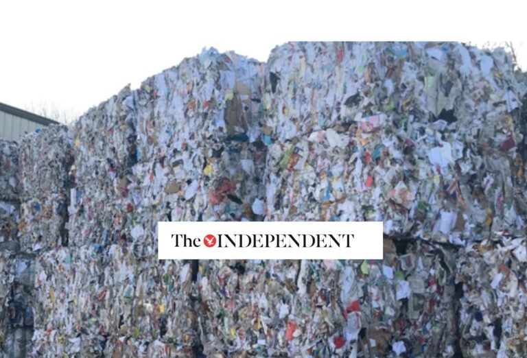Large stacks of compacted mixed paper and cardboard waste are piled outdoors. The logo of The Independent is overlaid at the center of the image.