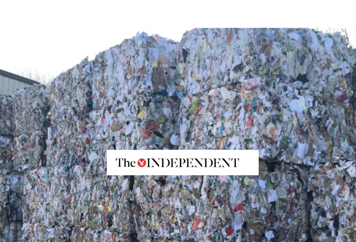 Large stacks of compacted mixed paper and cardboard waste are piled outdoors. The logo of The Independent is overlaid at the center of the image.