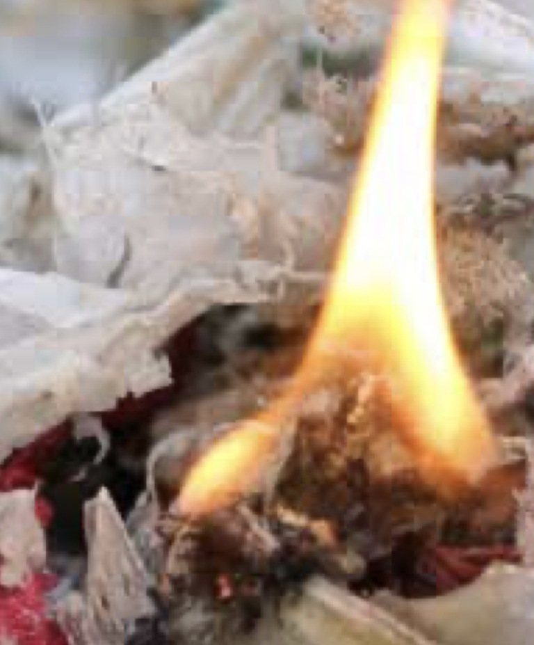 A close-up view of a small flame burning on a pile of crumpled paper and debris. The background appears out of focus.