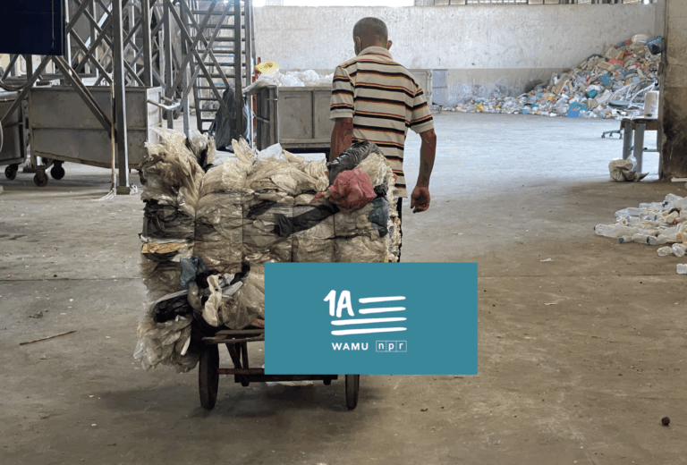 A man in a striped shirt pushes a cart loaded with plastic waste inside a recycling facility. Piles of plastic and industrial equipment are visible in the background. The 1A WAMU NPR logo is overlaid at the bottom.
