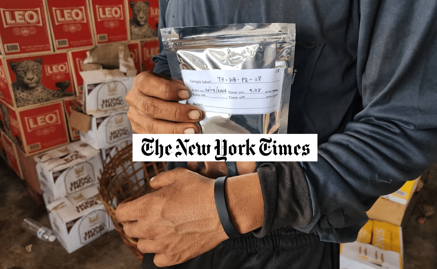 A person holds a sealed silver packet with a label, standing in front of stacked boxes and a wicker basket. The New York Times logo is superimposed over the image. The person wears a dark long sleeve shirt and wristband.