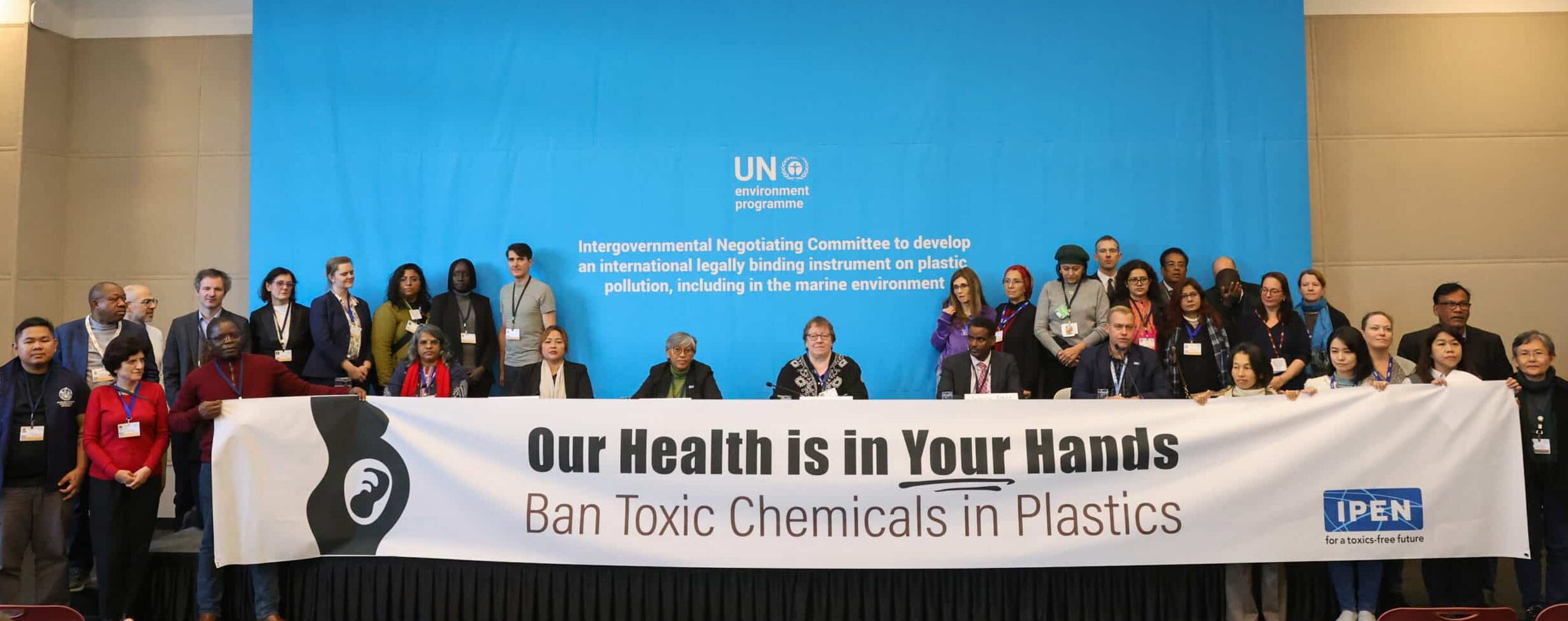A diverse group of people stands on a stage holding a large banner that reads, “Our Health is in Your Hands. Ban Toxic Chemicals in Plastics.” A blue United Nations Environment Programme backdrop is behind them.