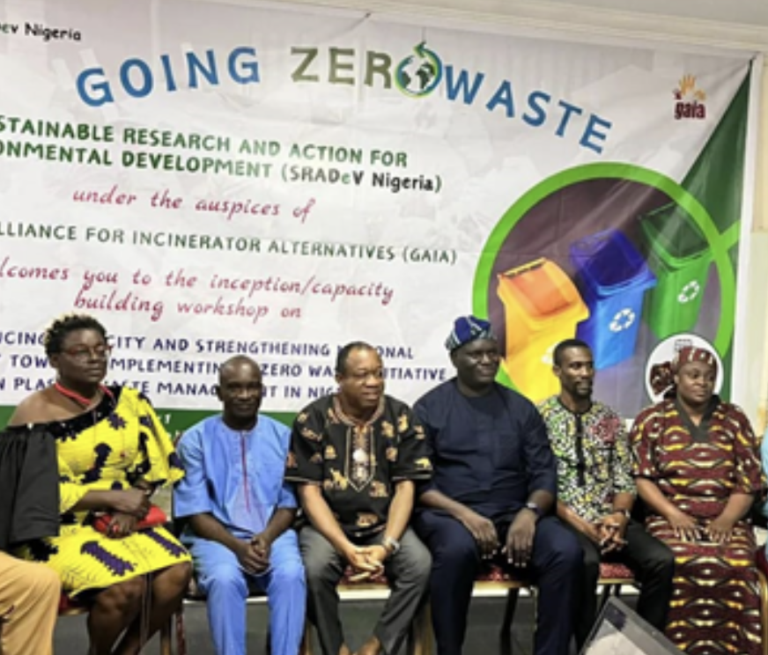 Seven adults sit side by side in front of a colorful “Going Zero Waste” banner at an environmental event. They face the camera, with recycling icons and text visible on the backdrop behind them.