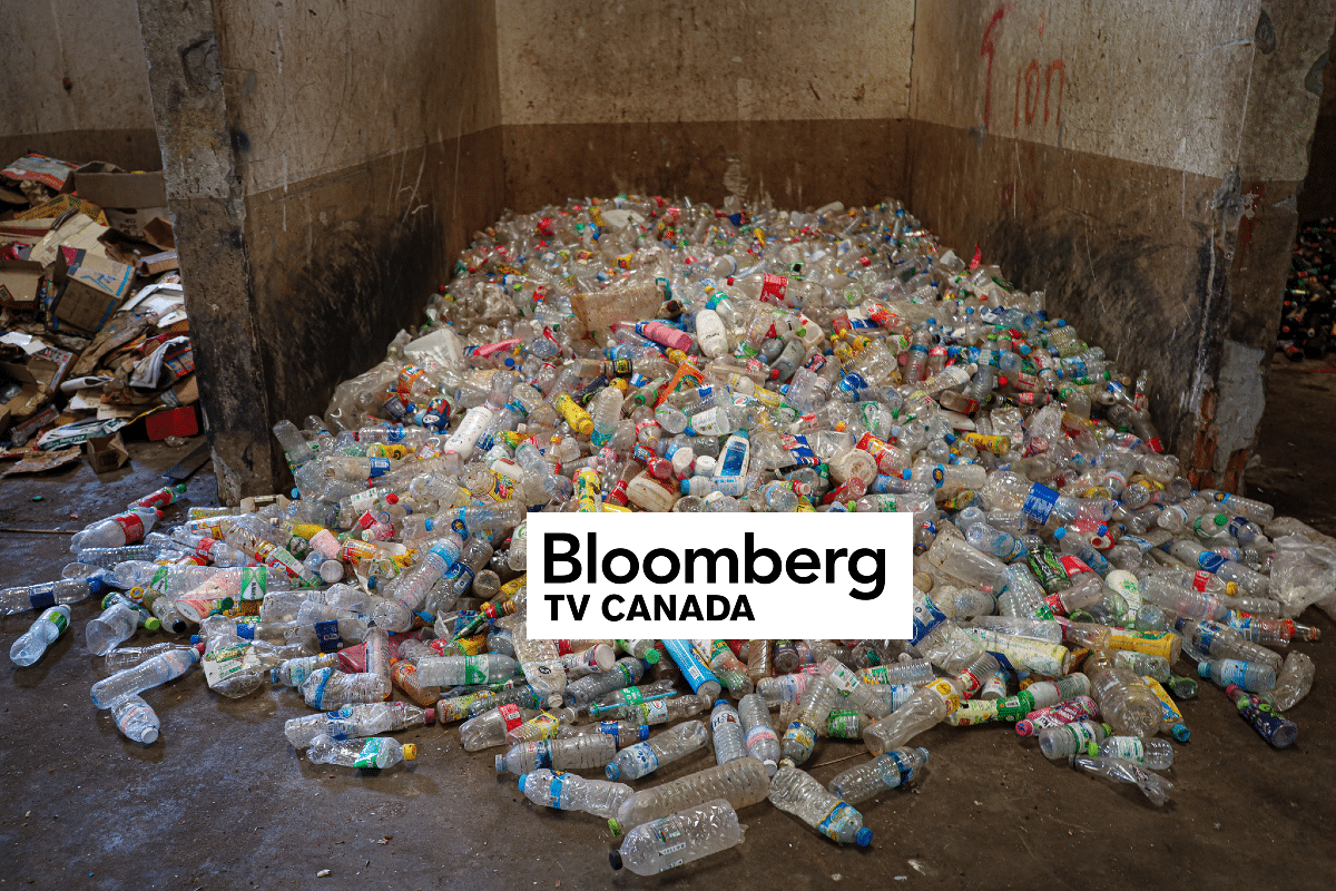 A large pile of empty plastic bottles covers the floor of a dirty, concrete room. Cardboard and more debris are seen in the background. A Bloomberg TV Canada logo is superimposed over the image.