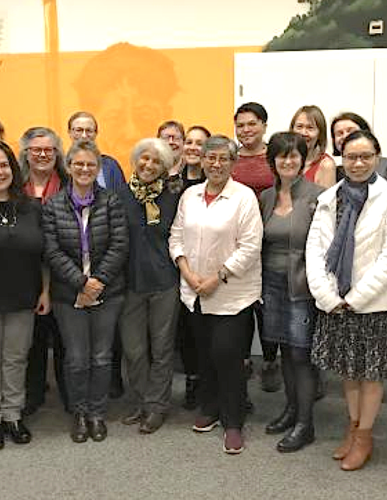 A group of fifteen adults, standing close together indoors, pose for a photo. Most are smiling. The background includes an orange wall with artwork and a white door.