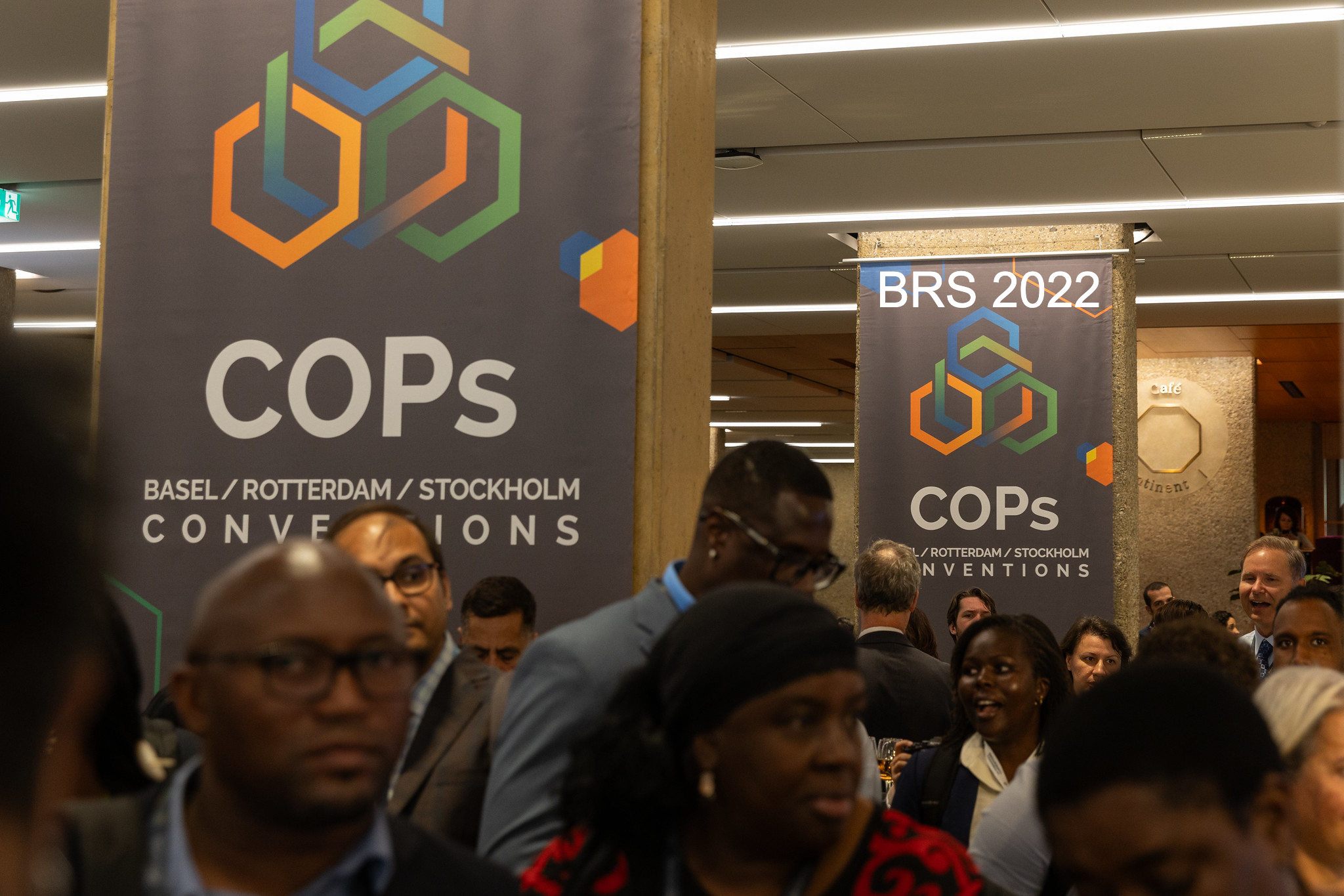 A crowd of people gather indoors in front of large banners reading COPs Basel / Rotterdam / Stockholm Conventions and BRS 2022 at an event, with overhead lighting and a concrete pillar visible.