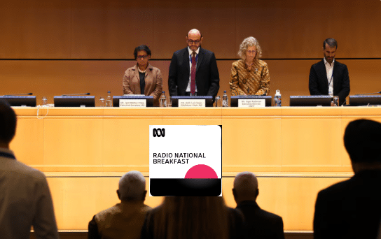 Four people stand behind a long panel table on a stage, facing an audience. Nameplates and microphones are in front of them. An overlay for “Radio National Breakfast” appears in the center foreground.