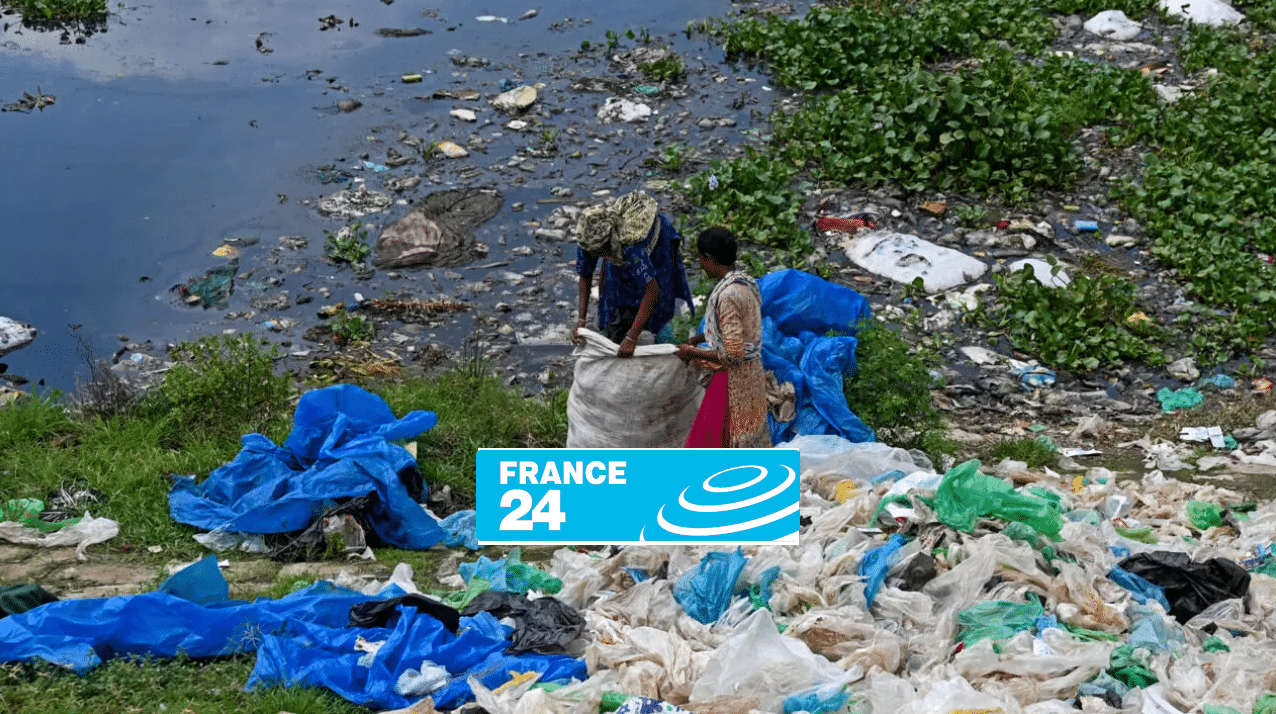 Two people collect plastic waste and garbage near a polluted body of water, with scattered debris and blue tarps on the grass. The France 24 logo is partially covering the lower center of the image.