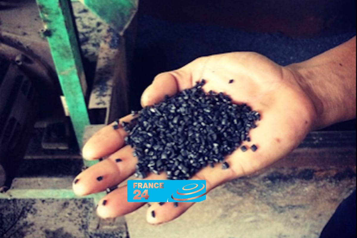 A hand holds a pile of small black plastic pellets; machinery and work surfaces are visible in the background. There is a France 24 logo at the bottom of the image.