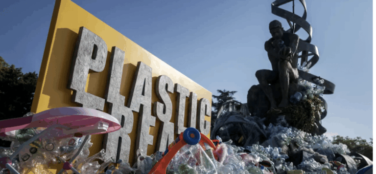 A large yellow sign reading PLASTIC is surrounded by a pile of plastic waste. In the background, a statue resembling Rodins The Thinker sits on top of more plastic, with a DNA helix structure behind it.