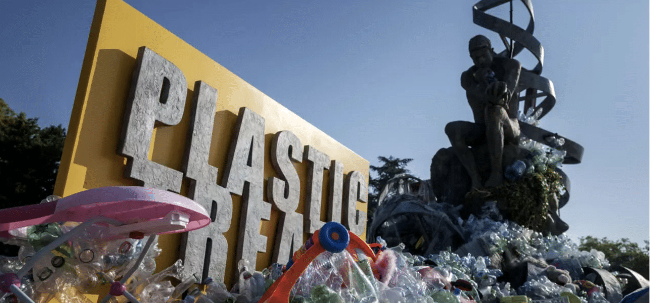 A large yellow sign reading PLASTIC is surrounded by a pile of plastic waste. In the background, a statue resembling Rodins The Thinker sits on top of more plastic, with a DNA helix structure behind it.