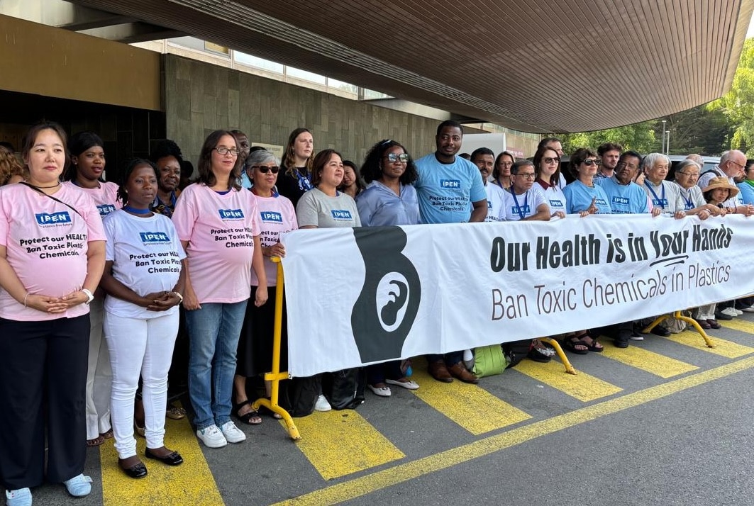 A group of people stands outdoors holding a large banner that reads “Our Health is in Your Hands: Ban Toxic Chemicals in Plastics.” Some people wear pink or blue shirts with similar messages.