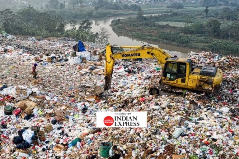 A yellow Komatsu excavator sits on a large landfill filled with scattered waste and plastic. A person walks nearby. In the background, there is a river and dense green vegetation. The Indian Express logo is visible in the middle.