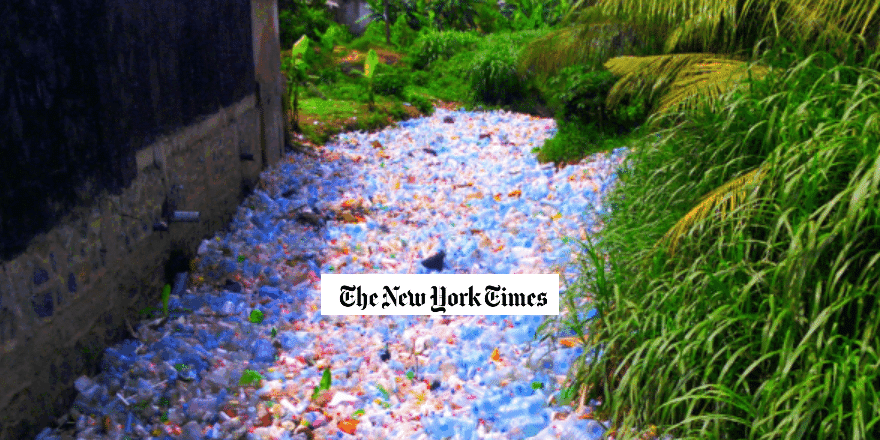 A narrow waterway is filled with plastic bottles and other litter, surrounded by dense green vegetation and a building wall. The New York Times logo is overlaid on the center of the image.