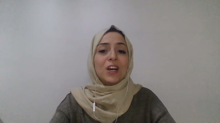 A woman wearing a beige hijab and a dark top is speaking. She has a wired earbud in her left ear and is seated in front of a plain white wall.